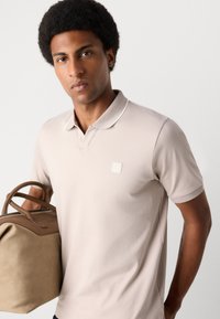 Beige polo ing galléros kivitelben, kis logóval ellátott folttal. A modell egy világosbarna táskát tart, amelynek formázott füllei vannak.
