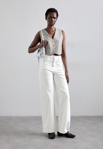 Un gilet gris court avec des boutons, associé à un pantalon blanc à jambes larges et des chaussures plates noires. Accessoirisé avec un petit sac à franges en argent.