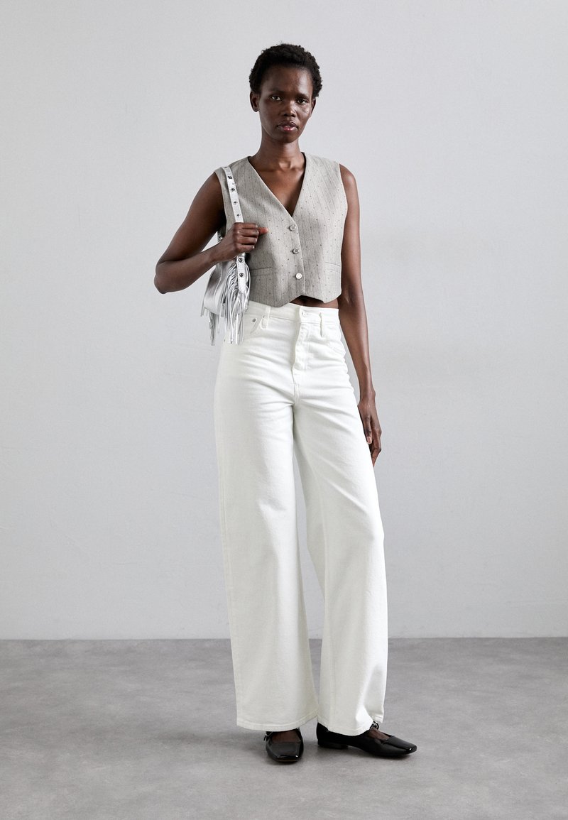 Un gilet gris court avec des boutons, associé à un pantalon blanc à jambes larges et des chaussures plates noires. Accessoirisé avec un petit sac à franges en argent.