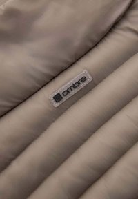 Details van een beige pufferjack met een gequiltte textuur, glad synthetisch materiaal en een duidelijk label met het zwarte "ombre"-logo.