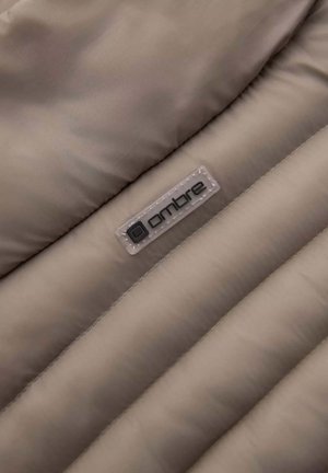 Détail d'une veste puffer beige mettant en valeur une texture matelassée, un matériau synthétique lisse et une étiquette claire avec le logo "ombre" en noir.