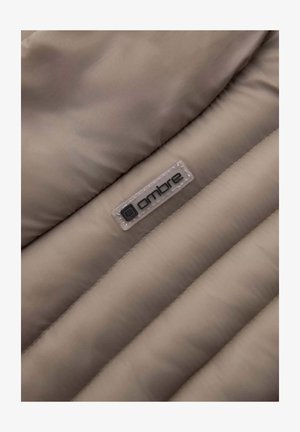 Detail eines cremfarbenen Daunenjacke, das die gesteppten Textur, das glatte synthetische Material und ein klares Etikett mit schwarzem "ombre"-Logo zeigt.