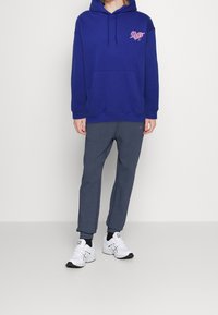 Tmavě modrá mikina s kapucí s růžovým potiskem na hrudi, doplněná šedými tepláky a bílými sportovními botami. Hladká tkanina, volný střih.