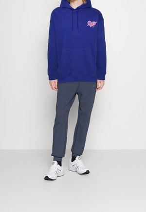 Felpa con cappuccio blu navy con grafica rosa sul petto, abbinata a pantaloni jogger grigi e scarpe da ginnastica bianche. Tessuto liscio, vestibilità casual.