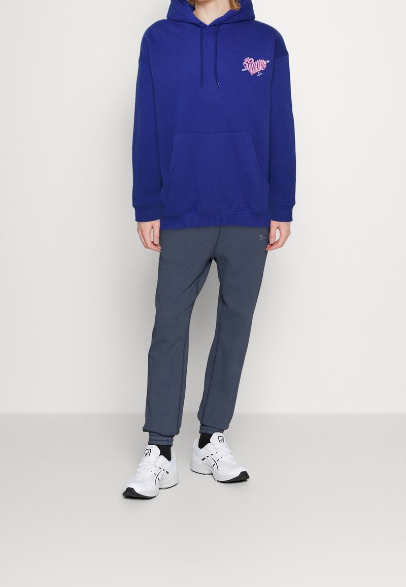 Tmavě modrá mikina s kapucí s růžovým potiskem na hrudi, doplněná šedými tepláky a bílými sportovními botami. Hladká tkanina, volný střih.