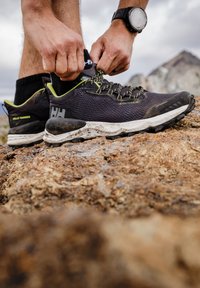 Černé běžecké boty do terénu s zelenými akcenty, texturední síťovinový svršek a bílá, zašpiněná mezipodešev. Osoba utahuje tkaničky na kamenitém terénu.