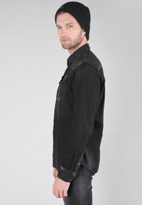 Camicia di jeans nera con design a bottoni, due tasche sul petto e maniche lunghe. Indossata con un berretto a coste nero.