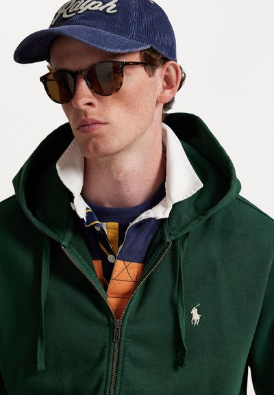 Homem vestindo um hoodie verde escuro com fecho, camisa branca de colarinho, camiseta de rugby listrada, boné azul com "Ralph" e óculos de sol com padrão de tartaruga.