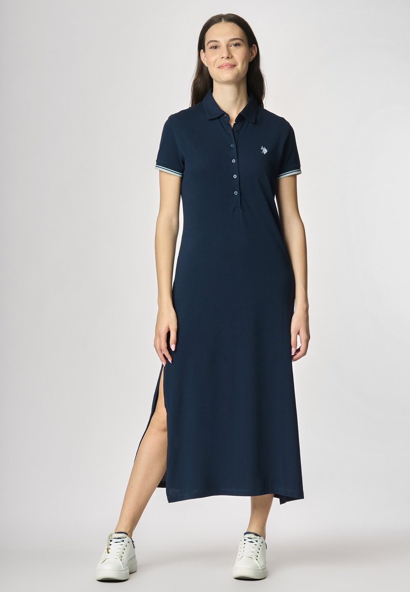 Abito polo navy con maniche corte, pattina dei bottoni, spacco laterale e accenti a strisce contrastanti. Indossato con sneakers bianche.