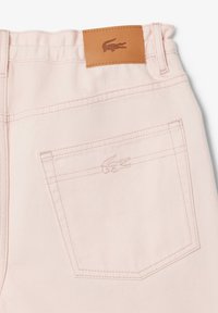 Ljust rosa denimbyxor har en texturerad yta, bakfickor med stickningar och en läderlapp med en logotyp på midjan.