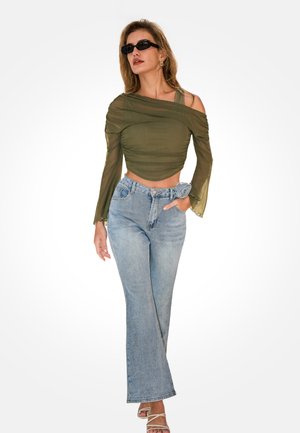 Femme portant un haut en maille asymétrique vert olive, un jean bleu clair avec une ceinture à fleurs en tissu, des lunettes de soleil et des talons blancs à brides, posant avec assurance.
