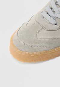 Svijetlosiva tenisica od brušene kože s teksturiranom površinom, ravnim vezicama i debelim potplatom u boji gume. Šavovi duž rubova.