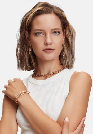 Collier en chaîne en or rose, bracelet assorti et boucles d'oreilles ; les maillons sont ovales et imbriqués, texture lisse, porté sur un haut à côtes blanc.