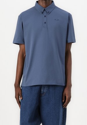 Mann trägt ein mattblaues kurzärmeliges Polohemd mit drei Knöpfen und einem gestickten Logo auf der linken Brust, kombiniert mit blauen Jeans.