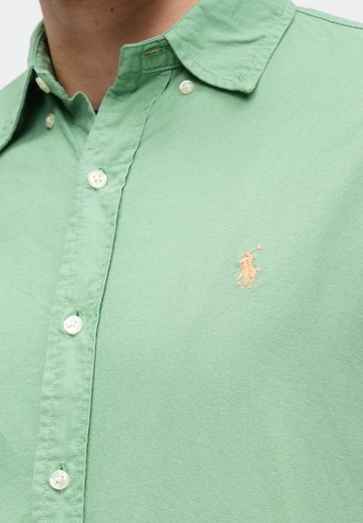 Homem a usar uma camisa verde-claro de botões com um pequeno logótipo bordado de jogador de polo no peito.