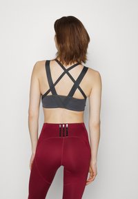 Sutiã desportivo cinza com alças cruzadas, apresentando uma textura de tecido suave. Combinado com leggings excessivamente altas em borgonha com detalhes em preto.