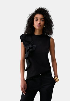 Donna con capelli ricci che indossa un top nero senza maniche con dettagli volant su una spalla e pantaloni neri, accessoriata con braccialetti d'oro e orecchini.