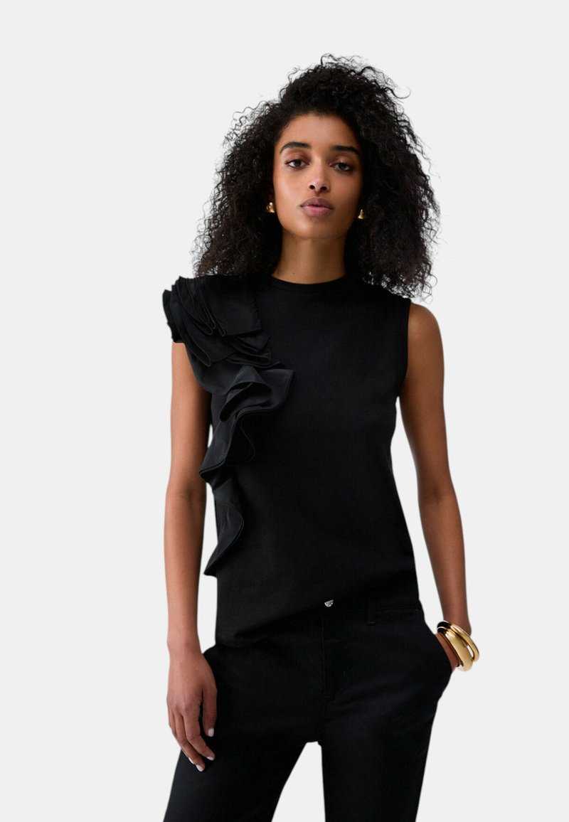 Femme aux cheveux bouclés portant un haut noir sans manches avec des volants sur une épaule et un pantalon noir, accessoirisée avec des bracelets et des boucles d'oreilles dorés.