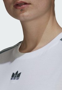 Balta džemperis su raukšlėta apykakle, su tamsiai žalia ir mėlyna siuvinėta logotipu ant krūtinės ir kontrastingomis juostelėmis ant pečių.