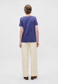 Μωβ κοντομάνικη μπλούζα με στρογγυλή λαιμόκοψη και μια μικρή λεπτομέρεια κουμπιού στην πλάτη, συνδυασμένη με κρεμ παντελόνι σε ευρεία γραμμή με ψηλή μέση.