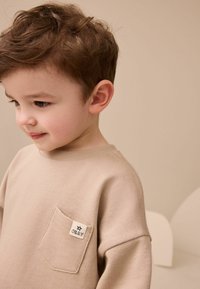 Beige sweatshirt med rund halsringning, korta ärmar och en framficka med en svart stjärnlogotyp. Mjukt, texturerat tyg.