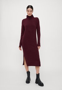 Burgundi turtleneck ruha kötött anyagból, hosszú ujjakkal és oldalsó hasítékkal. Fekete vastag bokacsizmával párosítva.