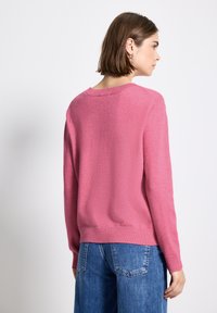 Donna con capelli corti castani, che indossa un maglione rosa e jeans blu, voltata di lato su uno sfondo bianco semplice.