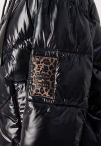 Sort pufferjakke med en blank finish, prydet med en leopardmønstret lapaccent med gyldne bogstaver. Letvægts, quiltet design for varme.