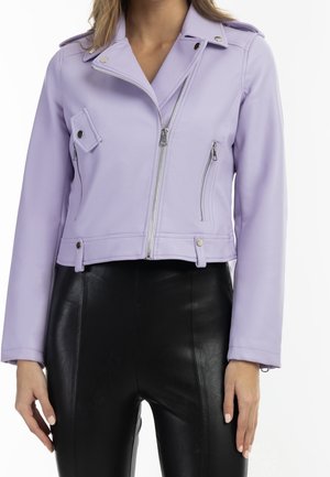 Femme portant une veste de motard violet clair zippée avec des boutons argentés et un pantalon en cuir noir taille haute, debout les bras détendus.