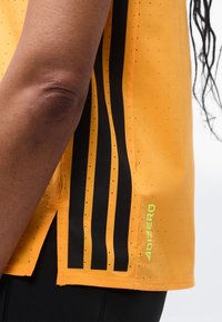 Κοντινό πλάνο ενός ατόμου που φοράει πορτοκαλί αθλητική μπλούζα με μαύρες ρίγες και κείμενο «ADIZERO» κοντά στο τελείωμα.