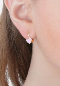 Orecchino d'oro con un piccolo ciondolo a forma di cuore rosa indossato all'orecchio sinistro di una persona con pelle chiara e capelli castani.