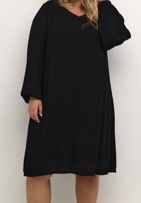 Robe noire longue jusqu'à la cheville avec une coupe ample, dotée de manches longues, d'un décolleté en V et de trois boutons au niveau du col, fabriquée en tissu texturé.