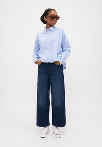 Šviesiai mėlyna marškinių tipo palaidinė, tamsiai mėlyni plačios kojų džinsai, balti sportbačiai ir tamsūs akiniai nuo saulės. Apranga turi laisvą siluetą ir neformalų įvaizdį.