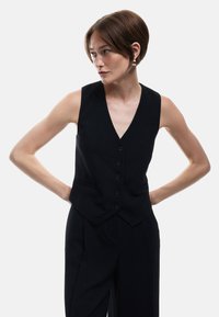 Zwarte mouwloze vest met een V-hals, voorzien van vijf knopen, twee zijzakken en een gestructureerde pasvorm, gecombineerd met wijde broekspijpen.
