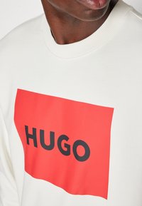 Felpa bianca con un grande patch rettangolare rosso con "HUGO" in bold nero. Collo a girocollo a coste e texture morbida.
