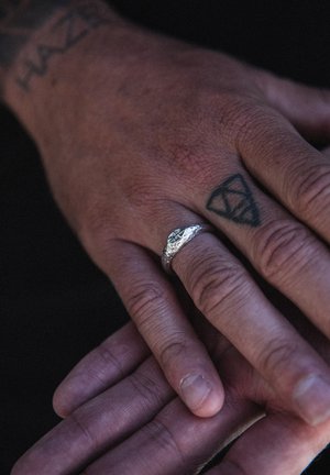 Bague texturée en argent sur une main brune, ornée d'un design rugueux. Une autre main se superpose, mettant en évidence un contraste de teinte de peau.