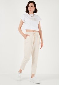 White cropped t-shirt with gold lettering, paired with beige joggers featuring an elastic waist and side pockets, complemented by white sneakers.