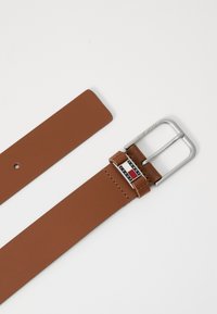 Brun lærbelte med en sølvfarget spenne, med logo i rødt, hvitt og blått. Glatt tekstur og enkelt design.