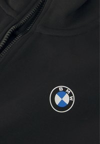 Haut de sweatshirt zippé noir avec une texture lisse, présentant un logo circulaire BMW en blanc et bleu sur la zone gauche de la poitrine.