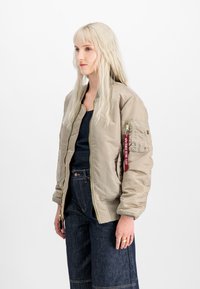 Beige bomber jacket with a zippered front, ribbed cuffs, and a patch pocket on the sleeve featuring a red tag. Smooth, shiny fabric finish.