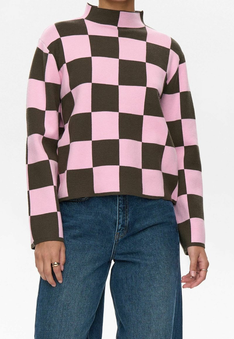 Personne portant un pull à carreaux rose et brun foncé à manches longues associé à un jean en denim bleu sur un fond uni.