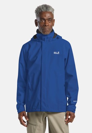 Homme âgé aux cheveux gris portant une veste imperméable bleue Jack Wolfskin et un pantalon beige, debout devant un fond uni.