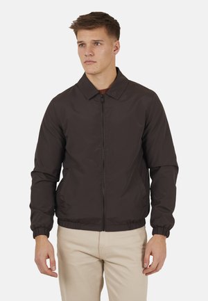 Młody mężczyzna w ciemnobrązowej kurtce z zamkiem błyskawicznym i elastycznymi mankietami oraz jasnobeżowych spodniach, stojący na jednolitym jasnym tle.