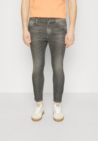Jeans gris ajustados con una textura deslavada, cuatro bolsillos y cierre de cremallera, combinados con zapatillas blancas y una camisa naranja.