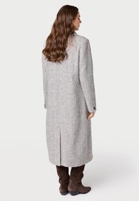 Cappotto in lana a spina di pesce grigio con una linea lunga e dritta, spacco laterale e polsini con bottoni. Indossato con stivali marroni al ginocchio.