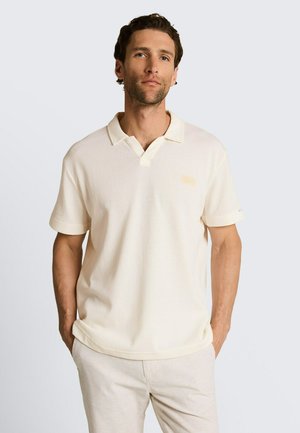 Mężczyzna stojący z rękami w kieszeniach, ubrany w kremową koszulkę polo z krótkim rękawem i jasnobeżowe spodnie na jednolitym tle.