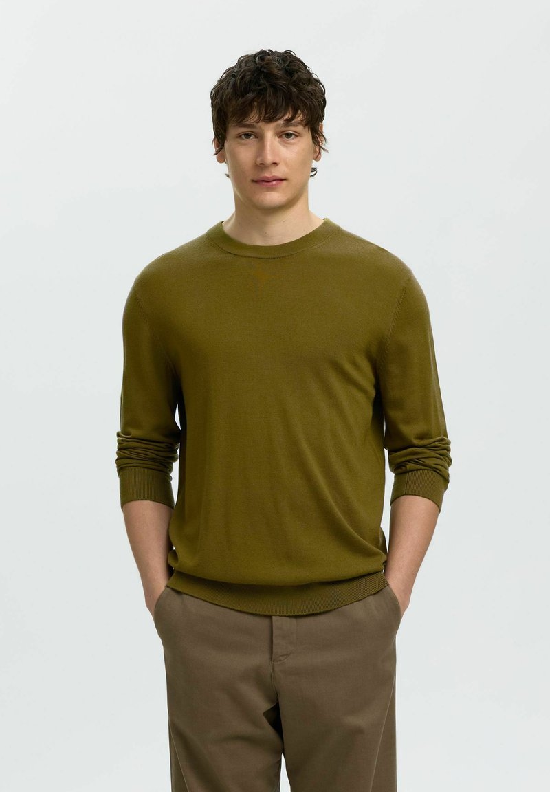 Jeune homme aux cheveux bouclés foncés portant un pull vert olive et un pantalon marron, debout les mains dans les poches devant un fond uni.