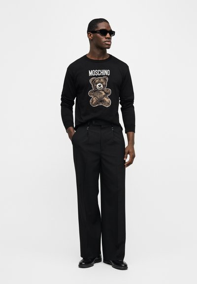 Άνδρας με μαύρα γυαλιά ηλίου, μαύρο μακρυμάνικο πουλόβερ Moschino με γραφικό αρκουδάκι, μαύρο παντελόνι φαρδύ και μαύρα παπούτσια, στέκεται μπροστά σε λευκό φόντο.