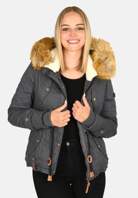 Smiling woman wearing a dark gray winter jacket with a tan fur-lined hood, holding the jacket collar over a black top and pants.