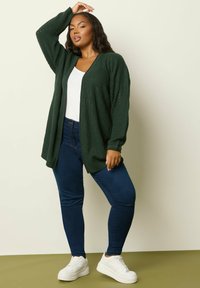 Mörkgrön stickad cardigan med öppen front och ballongsleeves, kombinerad med mörkblå tajta jeans och vita sneakers, framför en ljus bakgrund.